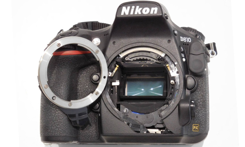 Nikon D810 de face montrant la cage miroir arrachée avec la bague de monture séparée du boîtier, exposant le capteur et les lamelles d'obturateur