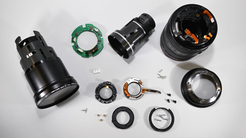 Objectif Canon démonté en ses éléments principaux : groupes optiques, barillet, circuit électronique, bague de contacts et visserie, étalés sur fond blanc