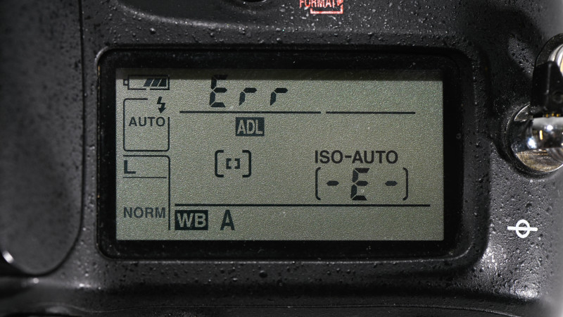 Gros plan sur l'écran de contrôle d'un Nikon affichant le message "Err" avec ISO-AUTO et indication (-E-), appareil recouvert de gouttes d'eau